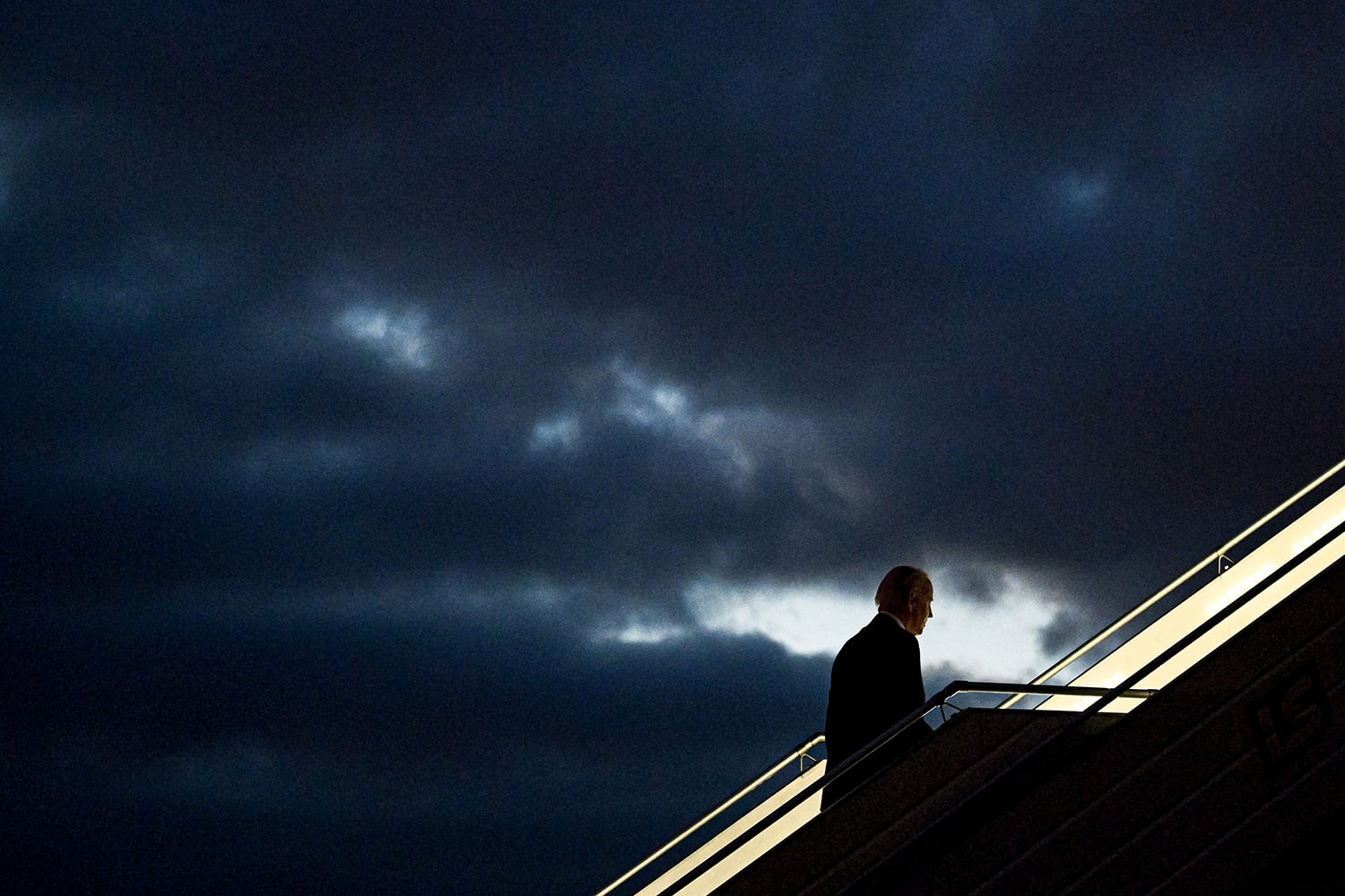 Joe Biden, in silhouette, walks up stairs to board a plane, with ominous dark clouds in the background.
