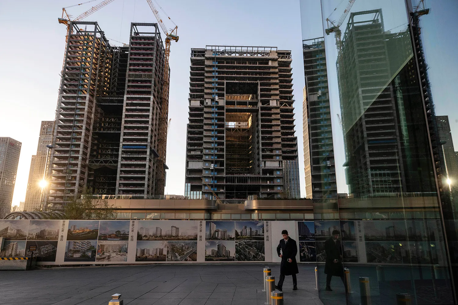 A man in a coat outside of a glass office building, with high rise buildings under construction in the background.