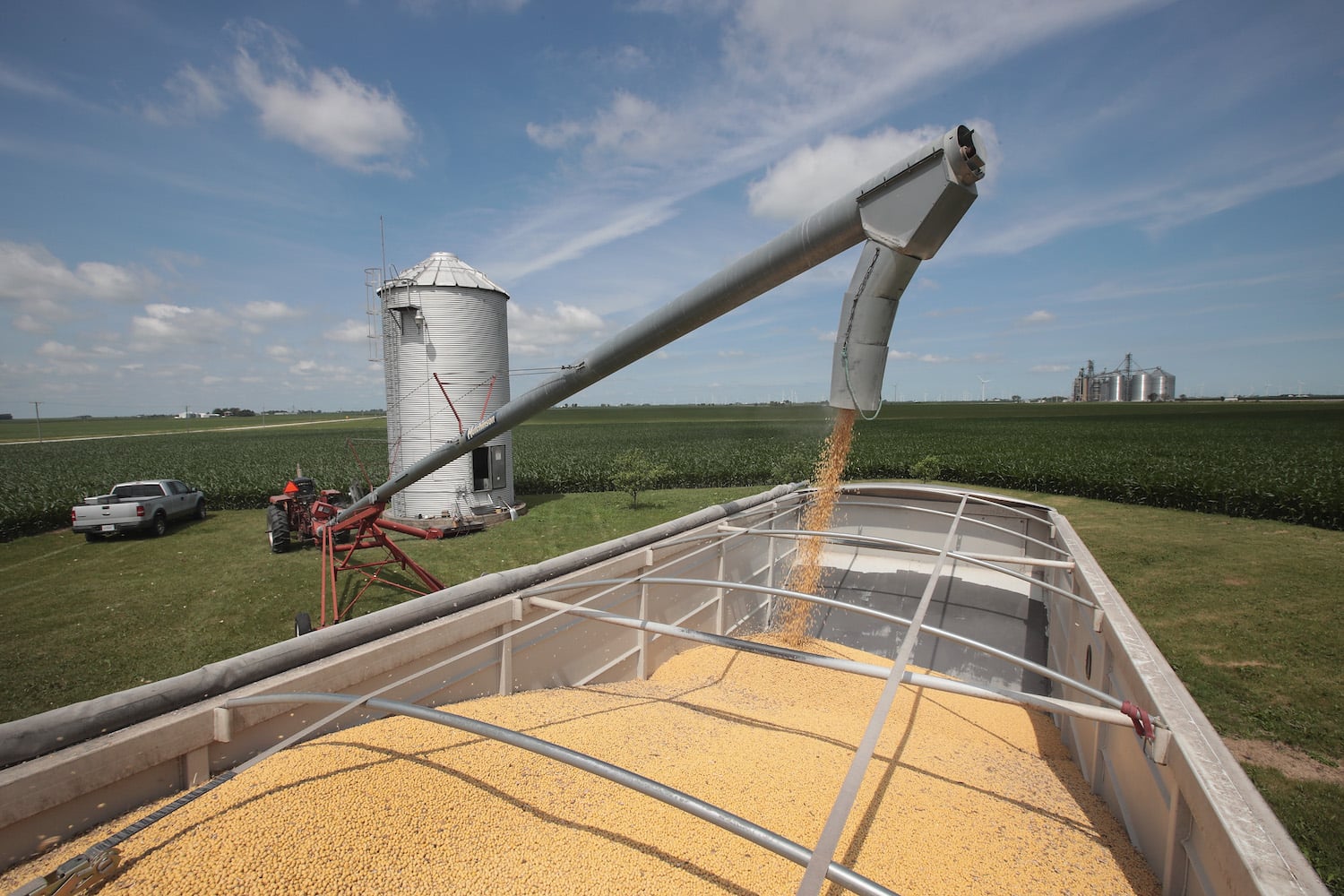A farmer uses the long, hollow arm of a machine to funnel soybeans from his small silo grain bin into the bed of a large truck. Lush green soybean fields stretch to the horizon in the distance beneath a partly cloudy sky.
