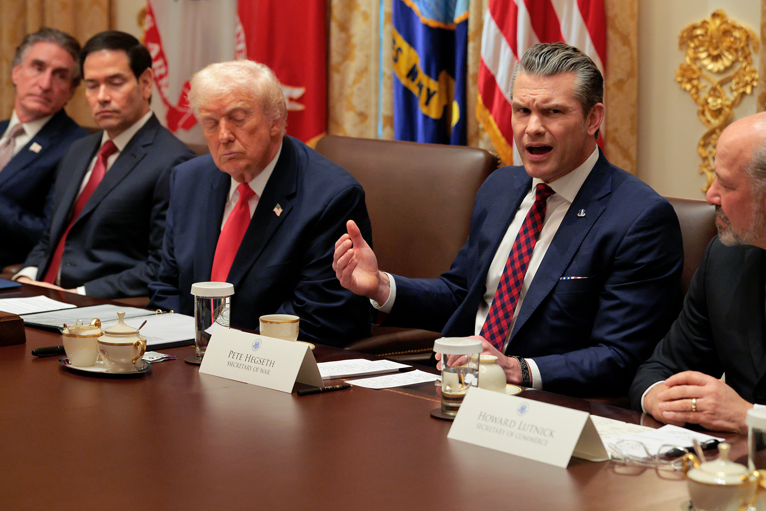 Trump, Hegseth, Rubio, and other officials sit at a table with papers and namecards in front of them. Hegseth has one hand raised and his mouth open as he speaks. Beside him, Trump is blinking and frowning.