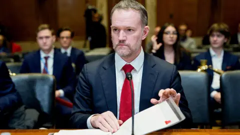 Getty Images Jamieson Greer speaking in Congress. He is seated before a microphone, flipping through a binder. He is wearing a black suit with a bright red tie. He has short hear and a grey and brown short beard.