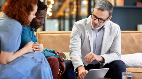 Getty Images Man and woman sit on a sofa where they are shown something on a tablet computer by a man in a blazer and shirt.