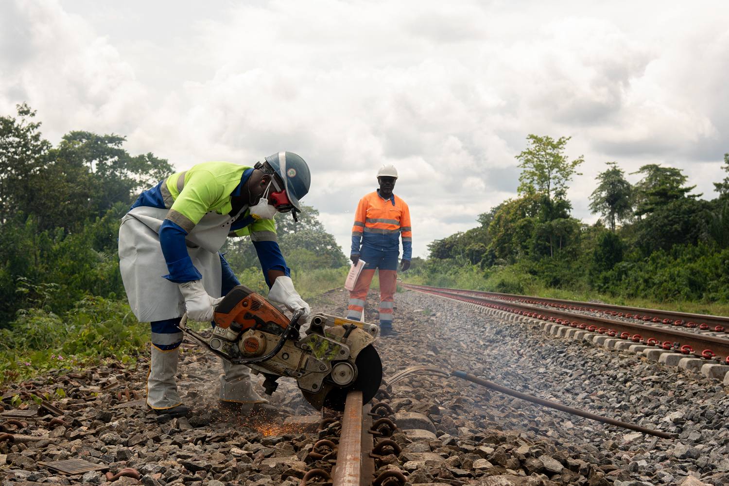 A man in the foreground bends over construction equipment he is using to renovate a section of railroad while his coworker looks on in the background in a isolated and forested area of Gabon.