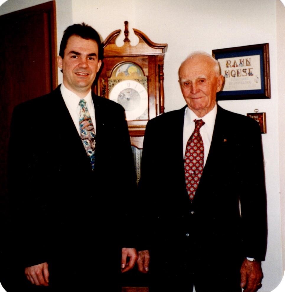An older man with thinning white hair stands next to a younger, taller man with dark hair. Both wear dark suits.