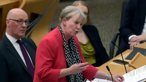 PA Media Finance Secretary Shona Robison announces the draft Budget for 2026-27 to MSPs at the Scottish Parliament in Holyrood. She stands in the debating chamber at a desk with two folders in front of her. She has short, tied back fair hair and wears a pink top and black and white scarf and is speaking. John Swinney sits next to her and looks on, as do other MSPs in the background.