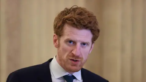PA Media Matthew O'Toole has red hair and a thin red beard and is wearing a dark suit jacket and tie and light shirt