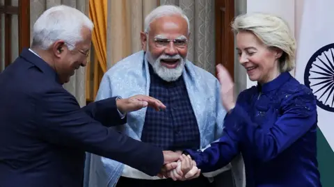 Reuters Three leaders are standing close together indoors, smiling and laughing during a friendly moment. The man in the centre, Indian Prime Minister Narendra Modi, with a white beard and glasses, wears a light shawl over a dark outfit and appears animated. On either side, European Council President António Luís Santos da Costa (left) in a dark suit and European Commission President Ursula von der Leyen (right) a deep blue jacket lean in, clasping hands in a gesture of warmth. A national flag and curtains are visible behind them