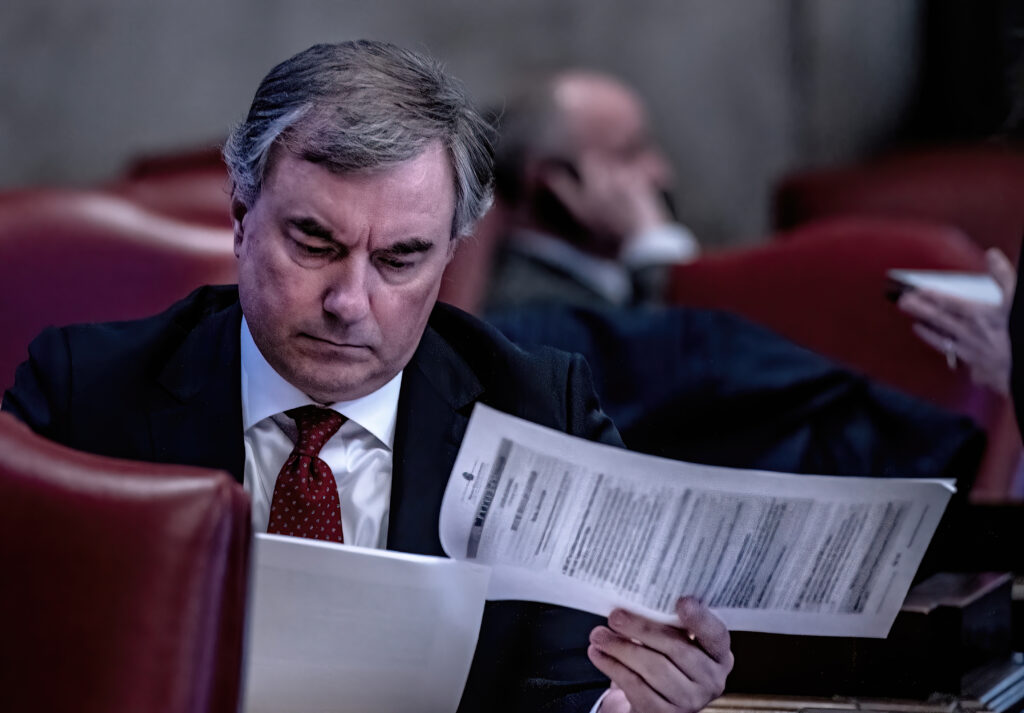 A man with gray hair tilts his head down to read a sheaf of documents.