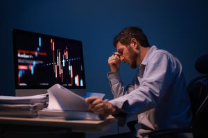 A man sitting at a desk analyzing financial data on a computer screen looking stressed while holding documents