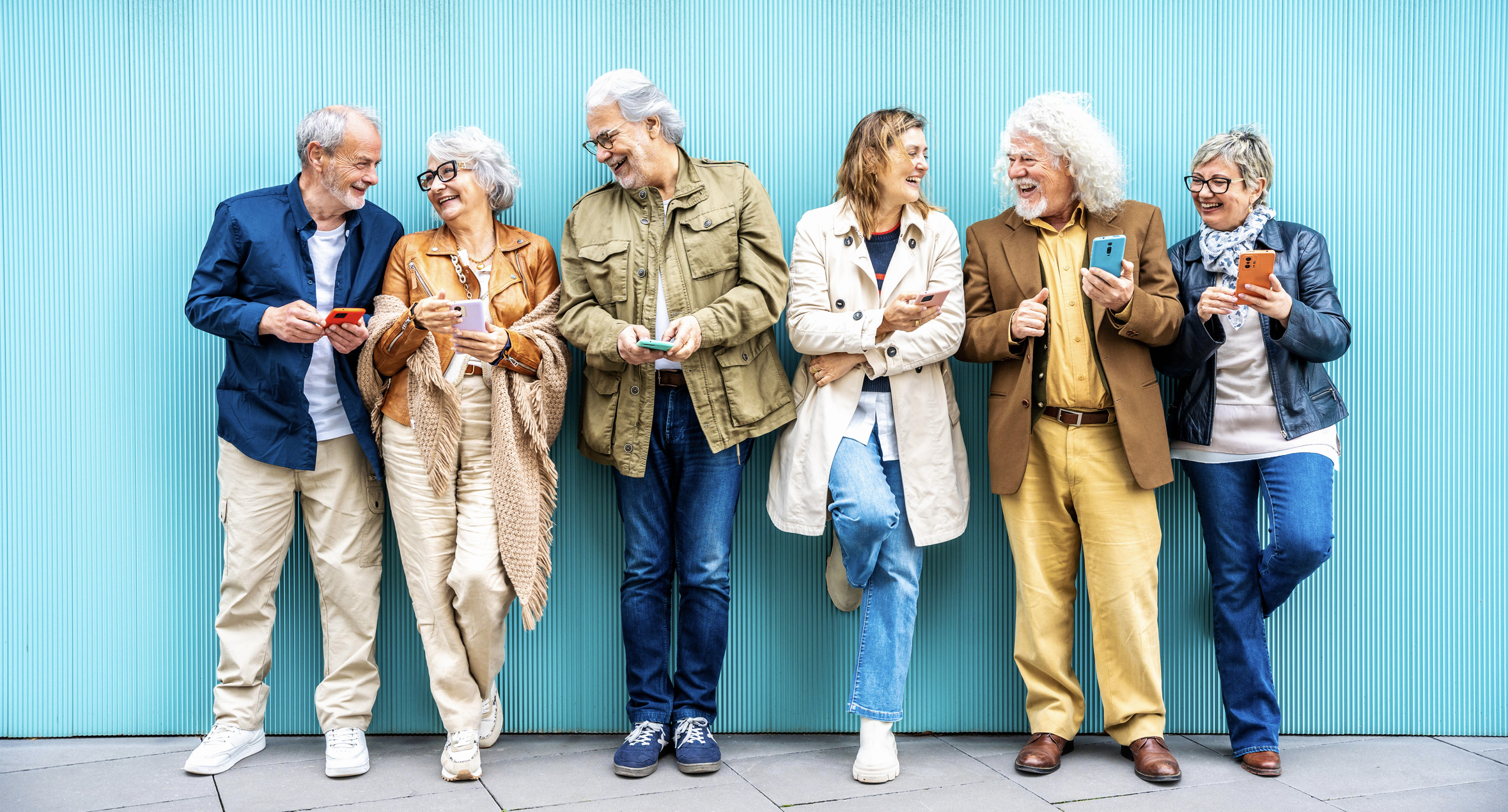 Group of senior people using technology devices together standing on a blue wall - Happy older friends having fun watching funny video on smartphone - Tech and modern elderly concept