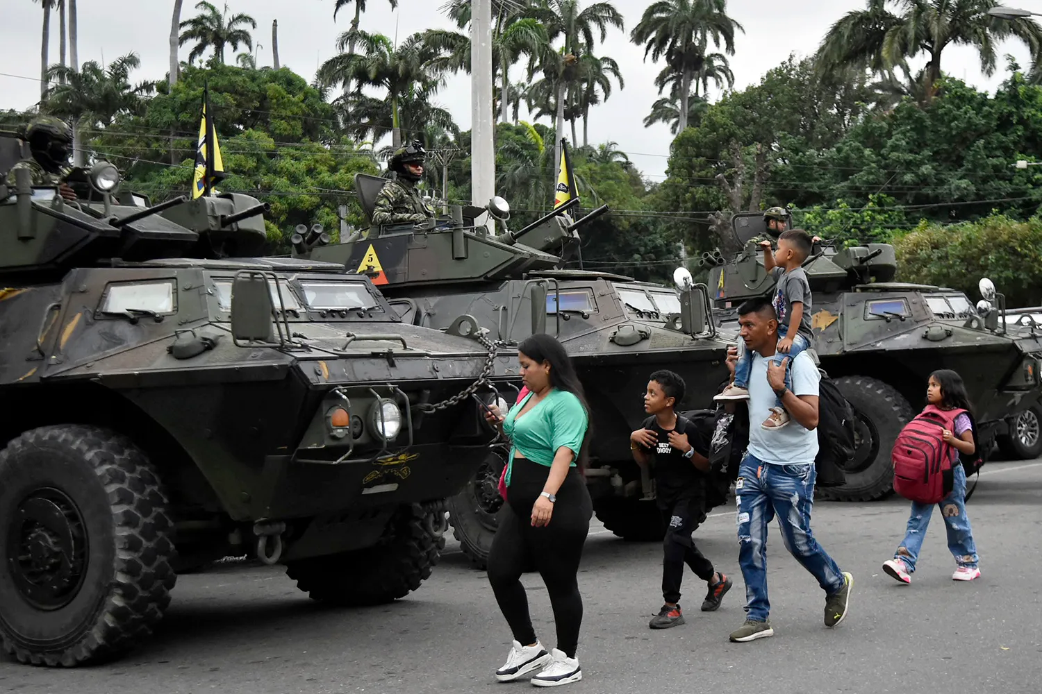 A woman and man walk with three children, one on the man's shoulders, past a row of armored military vehicles with soldiers in combat gear. The children carry backpacks. Palm trees are seen in the distance.