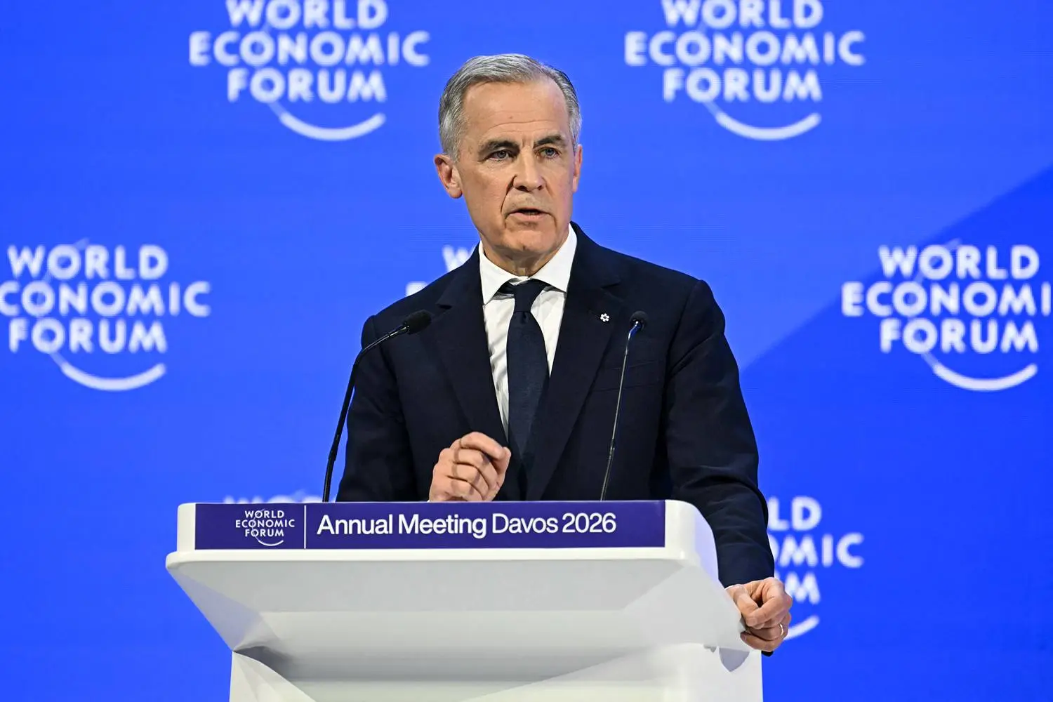 Carney stands at a lectern in front of a blue backdrop featuring the World Economic Forum logo. He wears a black business suit suit and tie.