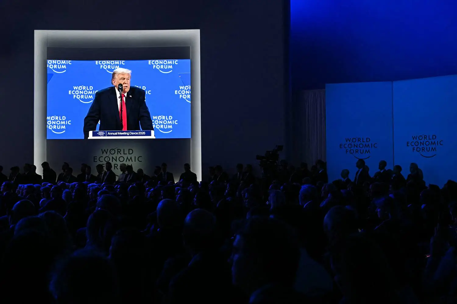 Trump is shown standing at a lectern on a large screen while the audience looks on in a darkened room.