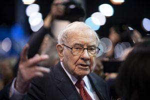 Warren Buffett in a public setting gesturing with his hand surrounded by people and cameras