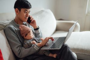 A person holding a baby and working on a laptop while speaking on a phone