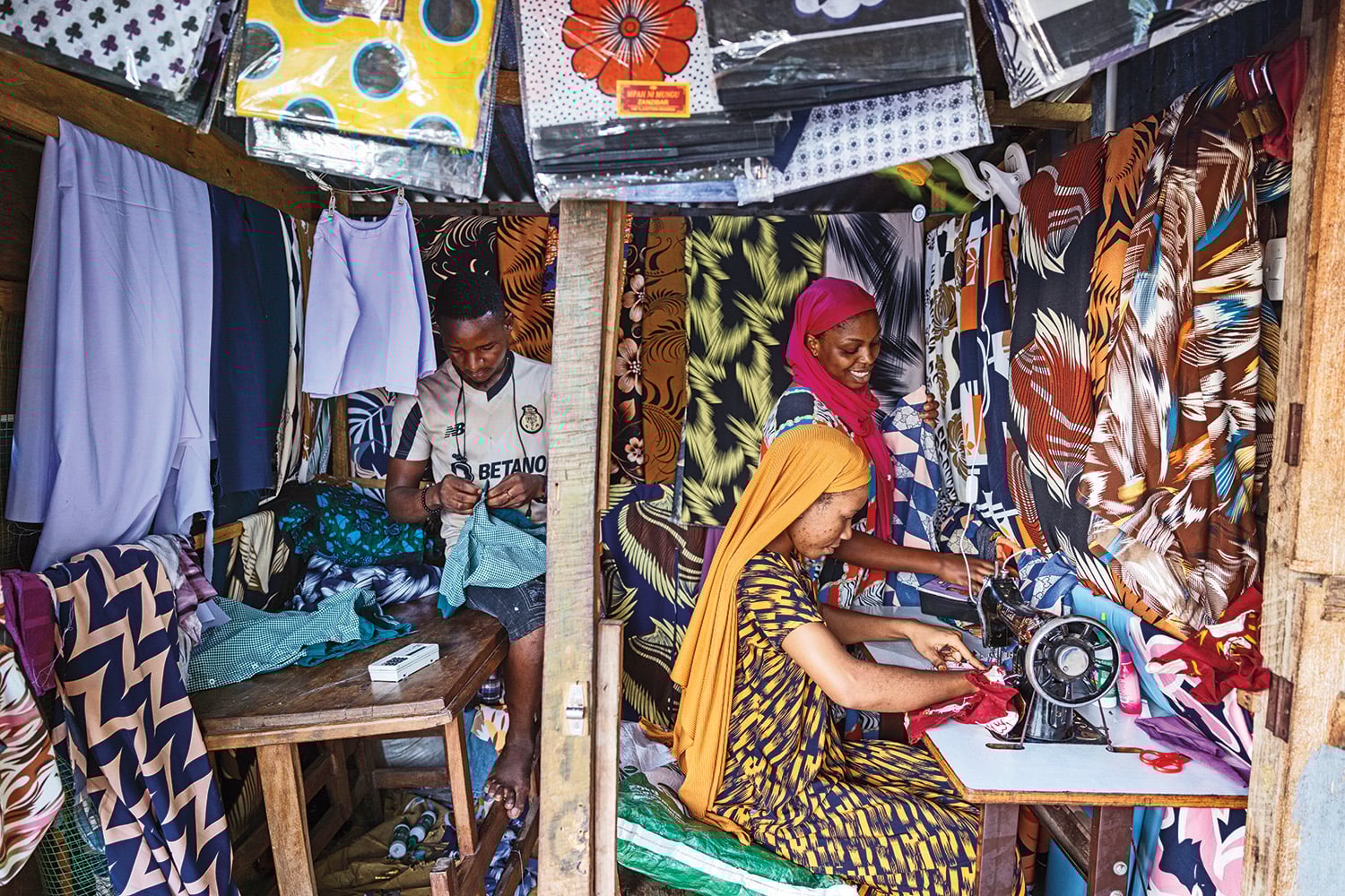 Two women work at a sewing machine in a booth filled with patterned fabrics. A man is seen working on fabric at a table on the left.