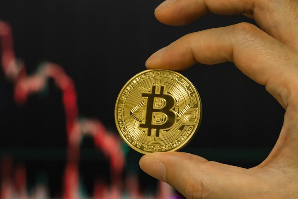 CHONGQING, CHINA - FEBRUARY 4: In this photo illustration, a person holds a Bitcoin coin in front of a blurred market chart showing a downward trend, on February 4, 2026, in Chongqing, China. Bitcoin fell to its lowest level since U.S. President Donald Trump returned to office, dropping below the $75,000 mark amid sustained selling pressure, as traders turned more cautious toward risk assets and options markets signaled limited appetite for a rebound, according to recent market data. (Photo illustration by Cheng Xin/Getty Images)
