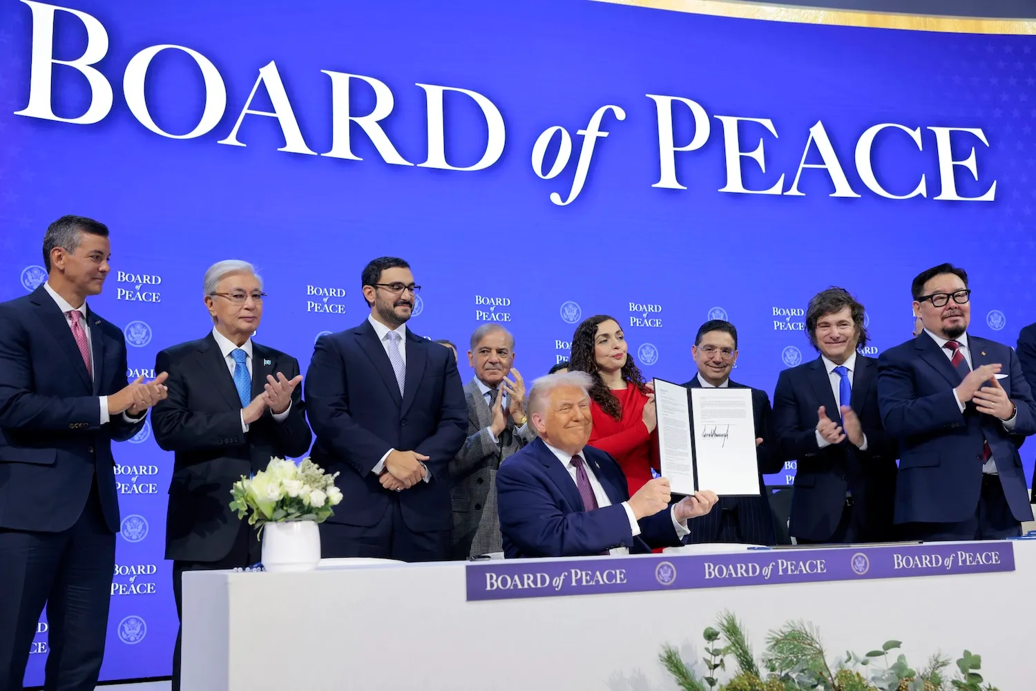 Trump beams and sits at a long table while holding up a folder containing printed text as well as his large signature. Other world leaders stand behind him, some with their hands clasped, some clapping.
